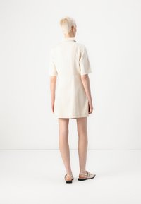 Robe en coton beige à manches courtes, col polo, et coupe décontractée, se terminant au-dessus des genoux. Portée avec des sandales grises claires.