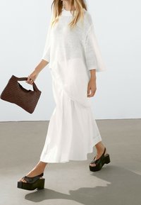 Witte gebreide oversized top, vloeiende witte rok en zwarte platform sandalen. Bruine geweven tas heeft een gestructureerd handvat.