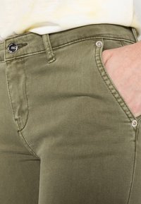 Jeans verde oliva de textura suave, con un bolsillo frontal, un botón de metal y costuras reforzadas en las uniones.