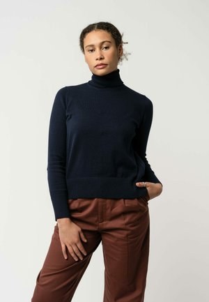 Navyblauer Rollkragensweater mit langen Ärmeln und geripptem Saum, kombiniert mit braunen hochgeschnittenen Hosen, die eine glatte Textur und eine maßgeschneiderte Passform aufweisen.