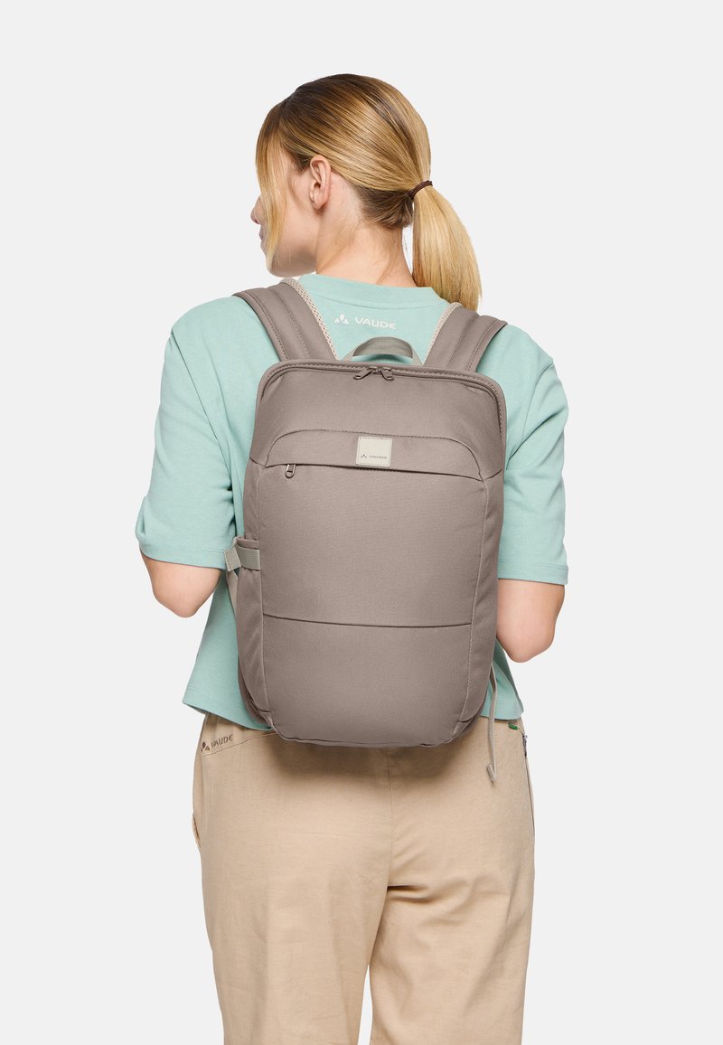 Persona con coda di cavallo bionda che indossa una maglietta verde menta e pantaloni beige, con uno zaino VAUDE taupe, vista da dietro.