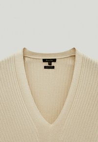 Beiger gerippter Strickpullover mit tiefem V-Ausschnitt, mit einem Etikett, auf dem "Made in Turkey" und Größenangaben stehen. Weiche Textur, leichtes Design.