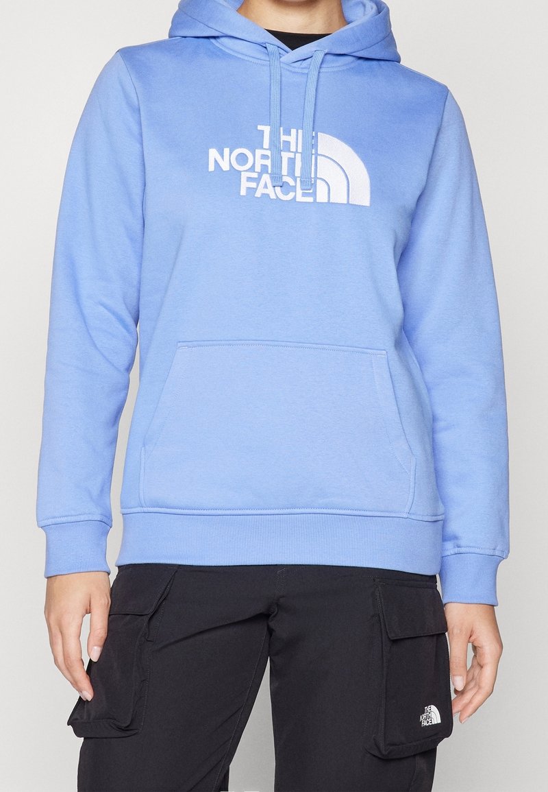 Personne portant un sweat à capuche bleu clair avec le logo "The North Face" et un pantalon cargo noir avec des poches, face à un fond uni.