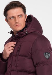 Giacca a vento bordeaux con cappuccio, tasche con cerniera, texture trapuntata e una toppa con logo sulla manica sinistra. Materiale morbido e isolante.
