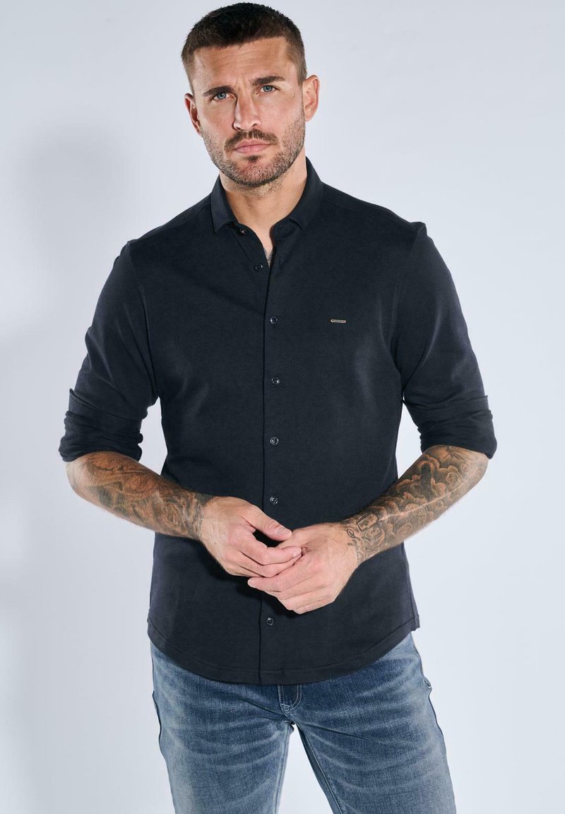 Schwarzes Langarmhemd mit Knopfverschluss, taillierte Passform und dezentem Logodetail auf der Brust. Getragen mit blauen Jeans, die die Textur der Ärmel hervorheben.