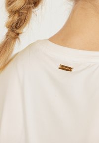 Valkoinen t-paita, jossa on sileä pinta, ja takana pieni kultainen merkki. Yksinkertainen muotoilu, jossa on pyöreä kaula-aukko.