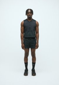 Schwarze Sportweste mit Reißverschluss, kombiniert mit schwarzen Shorts. Getragen mit schwarzen Nike-Socken und Turnschuhen. Slim Fit, leichtes Material, minimales Design.