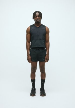 Schwarze Sportweste mit Reißverschluss, kombiniert mit schwarzen Shorts. Getragen mit schwarzen Nike-Socken und Turnschuhen. Slim Fit, leichtes Material, minimales Design.