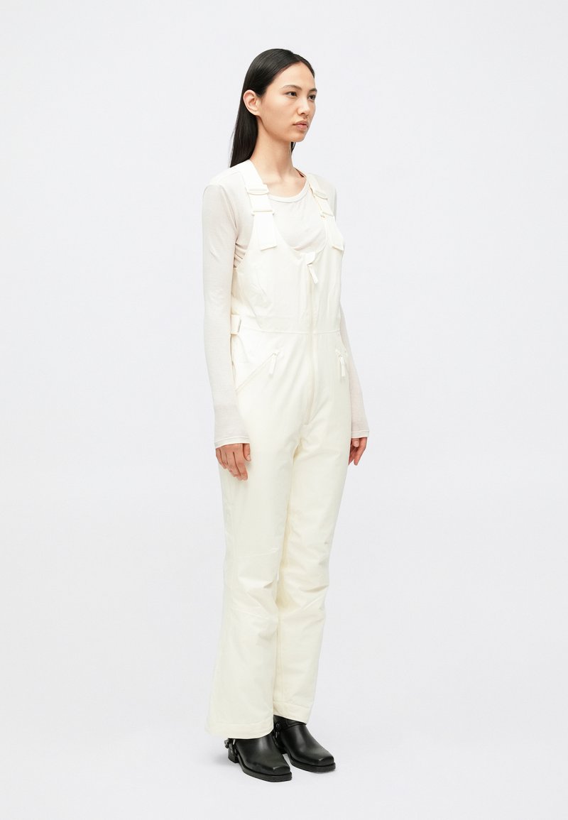 Tuta impermeabile bianca con bretelle regolabili, cerniera frontale, tasche laterali e gambe larghe, abbinata a stivaletti neri.