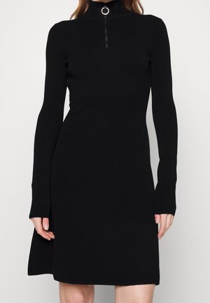 Schwarzes langärmeliges Strickkleid mit hohem Kragen und silbernem Ringreißverschluss-Detail, getragen von einer stehenden Person vor einfarbigem Hintergrund.