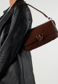 Brown leather handbag with a textured finish, gold hardware, and a heart-shaped clasp detail, worn on the shoulder against a black leather jacket.