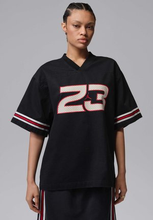 Jeune personne aux cheveux tressés portant un maillot de sport noir ample avec le numéro 23 et des accents de manches rayées rouge et blanc.