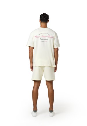 Άνδρας που φοράει κρεμ oversize T-shirt με ροζ κείμενο και ταιριαστά σορτς, στέκεται με την πλάτη στραμμένη μακριά σε λευκό φόντο.