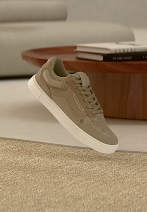 Beiger Freizeitschuh mit Mesh- und Velourslederdetails, weiße Sohle, schwebend über einem strukturierten beigen Teppich in einem modernen Wohnzimmer.