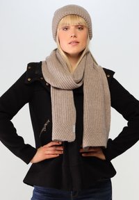 Beige strikket tørklæde og hat sæt, der har en ribbet tekstur. Tørklædet hænger elegant, mens hatten sidder tæt, begge lavet af blødt stof.