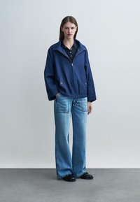 Frau steht vor einem einfarbigen Hintergrund und trägt eine marineblaue Reißverschlussjacke, ein schwarzes Hemd, weit geschnittene blaue Jeans mit Vorderentaschen und schwarze Schuhe.