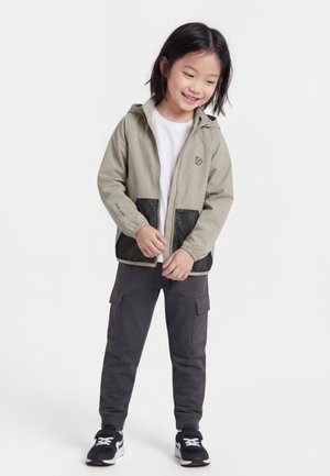 Malé dieťa oblečené v béžovej bunde s kapucňou a čiernymi sieťovinovými vreckami, bielej košeli, tmavých cargo nohaviciach a čiernych teniskách, stojace a usmievajúce sa.