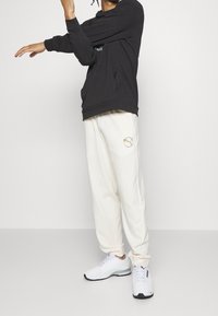 Person i svart hoodie och vita sweatpants som sträcker ut armen mot en enkel vit väggbakgrund.