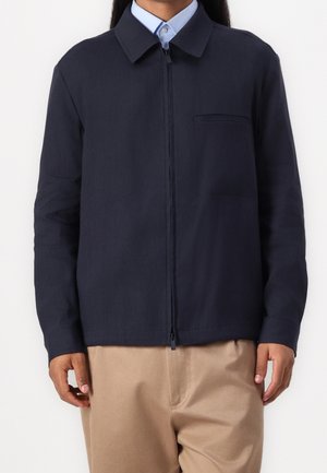 Homme portant une veste zippée bleu marine sur une chemise bleu clair à col et un pantalon beige, debout devant un fond uni.