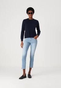 Navy hosszú ujjú pulóver, világoskék cropped farmernadrág oldalsó hasítékokkal és fekete slip-on cipő, viselve egy személy által, aki egy sima háttér előtt áll.