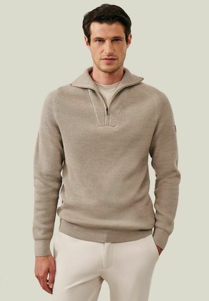 Man draagt een beige geribd trui met halve rits en licht crème broek met handen in de zakken tegen een effen lichte achtergrond.