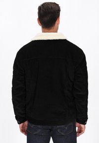 Schwarze Cordjacke mit einem weißen Schafslederkragen, ausgestattet mit geknöpften Manschetten und einem leicht übergroßen Schnitt. Minimalistisches Design ohne sichtbare Muster.