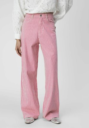 Kobieta ubrana w wysokie, różowo-białe spódnicowe spodnie w pionowe pasy, białą koronkową bluzkę i srebrne spiczaste buty, stojąca z jedną ręką na biodrze.