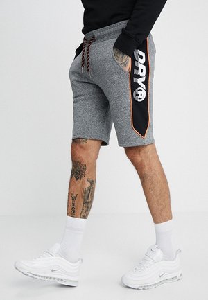 Mężczyzna w szarych szortach ze sznurkiem, z czarnymi i pomarańczowymi panelami po bokach oraz białych trampkach, pokazujący tatuaże na dolnych partiach nóg i dłoni.