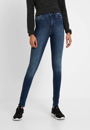Person trägt dunkelblaue Skinny-Jeans, schwarze Turnschuhe und einen dunkelgrauen Langarm-Pullover und steht mit leicht auseinanderstehenden Beinen.