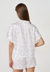 Vrouw in een witte korte mouwen pyjama met kleine roze, marineblauwe en bordeaux hartjespatronen, gezien van achter.