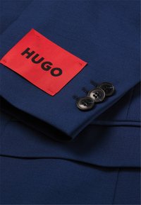 Navyblauer Anzugjackett mit einem roten rechteckigen Etikett mit der Aufschrift "HUGO" und drei schwarzen Knöpfen am Ärmel. Glatte Textur, maßgeschneiderte Passform.