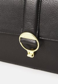 Μαύρη δερμάτινη τσάντα σε κοντινή λήψη, με χρυσό μεταλλικό κρίκο και ανάγλυφο λογότυπο, υφή στην επιφάνεια και προσεγμένες λεπτομέρειες ραπτικής.