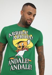 Vihreä puuvillapaita, jossa on sarjakuvahahmo, jolla on kellertävä hattu ja punainen solmio. Tekstissä lukee "ARRIBA! ANDALE!" lihavoiduin mustin kirjaimin.