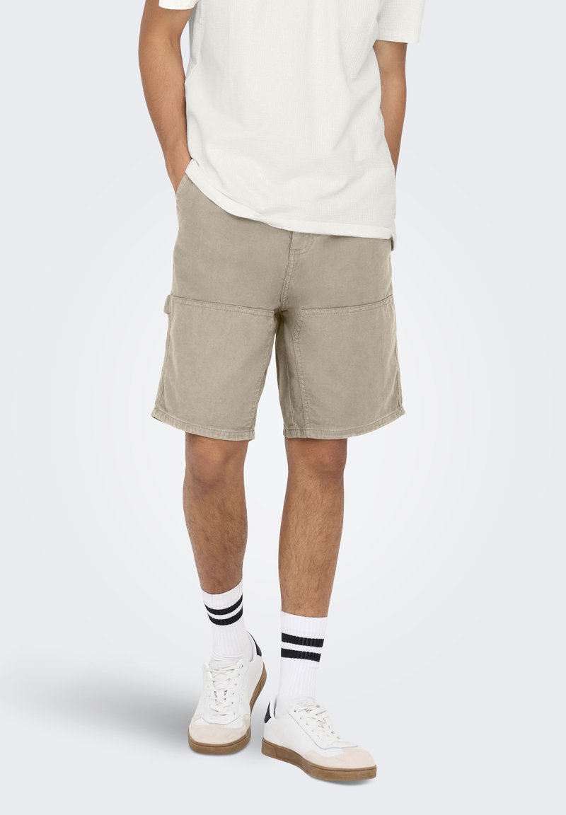 Pantalones cortos cargo caqui con una tela texturizada, que cuentan con múltiples bolsillos y un ajuste relajado. Combinados con calcetines blancos y zapatillas.