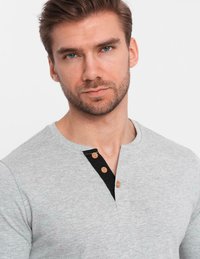 Ung man med kort brunt hår och skägg, klädd i en ljusgrå henley-tröja med tre träknappar och svart innerficka.