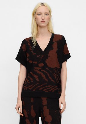 Blonde Frau trägt schwarze und braun gemusterte V-Ausschnitt-Strickoberteil mit kurzen Ärmeln und passende Strickhose vor einfachem hellem Hintergrund.