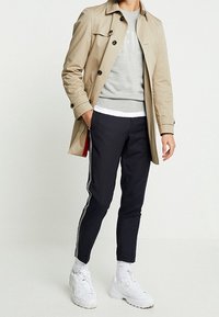 Trench coat beige sopra un maglione grigio, sottoveste bianca, pantaloni navy a gamba affusolata con strisce bianche e sneakers bianche. Tessuto liscio, vestibilità moderna.