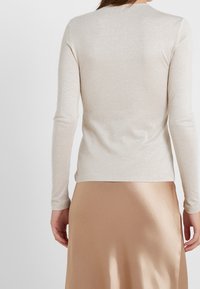 Beige pitkähihaine toppi kiiltävässä kankaassa, tiukasti istuvalla siluetilla ja pyöreällä kaula-aukolla, yhdistettynä sileään, neutraalin sävyiseen hameeseen.