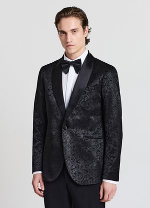 Giacca da smoking nera con intricati motivi floreali tono su tono, rever in raso a scialle, chiusura con un bottone, abbinata a una camicia bianca e papillon.