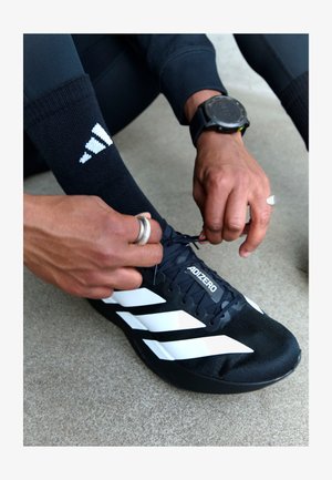 Pantof ca atletism de culoare neagră cu design în trei dungi albe, textură netedă și etichetă ADIZERO, fiind legat de un mâna care poartă inele.