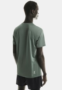 Tânărul îmbrăcat cu un tricou sport verde cu mânecă scurtă și pantaloni scurți negri, întors cu spatele, pe un fundal deschis neutru.