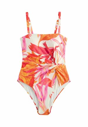 Jeune femme en maillot de bain fleuri sans bretelles debout à l'extérieur près d'un terrain rocheux et de verdure, regardant sur le côté avec les mains derrière le dos.