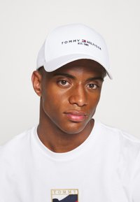 White baseball cap featuring the "TOMMY HILFIGER" logo in navy and red, worn by a person with a plain white t-shirt.