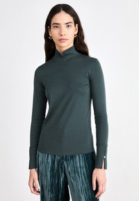 Dark green turtleneck top in stretchy fabric, featuring long sleeves with zippers at the cuffs, paired with pleated teal trousers.