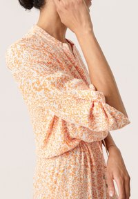 Šaty s květinovým vzorem v oranžové a bílé barvě, s manžetou na knoflíky a skladovanou texturou. Lehký materiál a uvolněný střih.