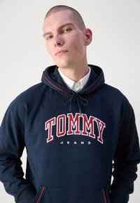 Námornícka modrá mikina s kapucňou vyrobená z bavlny. Obsahuje červené akcenty na šnúrkach, predné vrecko a veľké vyšiované "TOMMY JEANS" v bielej a červenej farbe.
