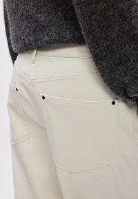 Παντελόνια σε κρεμ απόχρωση με λείο φινίρισμα, που διαθέτου�ν δύο πίσω τσέπες, μαύρες λεπτομέρειες από μεταλλικό υλικό και προσεγμένα ραμμένα ραψίματα.
