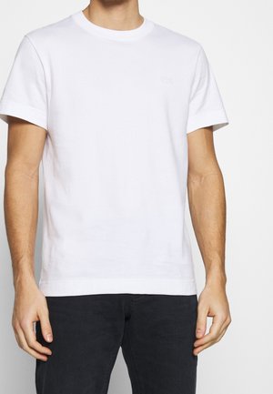 Man wearing plain white short-sleeve cotton T-shirt and black pants, standing against a white background with arms relaxed.