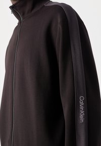 Gros plan d'une veste noire zippée avec une bande sombre sur la manche avec le texte du logo Calvin Klein en blanc.