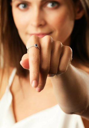 Frau zeigt einen silbernen Ring mit blauem Edelstein am Mittelfinger, trägt ein weißes Oberteil, unscharfer Hintergrund.